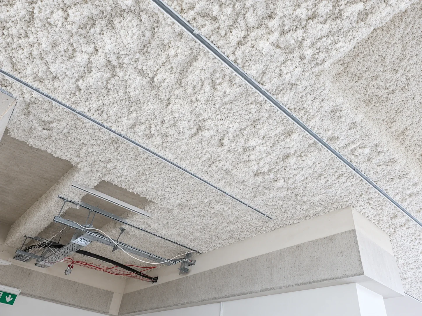 Sprayed acoustic finish across a concrete soffit and downstand, with lighting track and cable containment