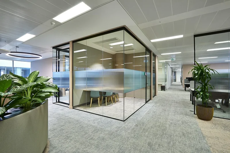 Glass-fronted meeting room with a fluted privacy band and pale timber trim, set in an open-plan office with planters and a ring pendant light