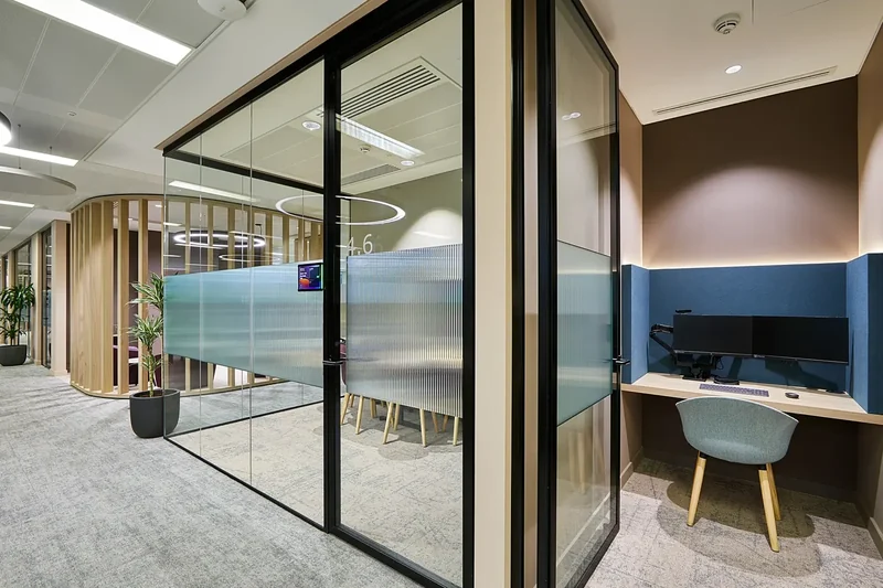 Glazed meeting room door beside a single-person focus booth with a blue acoustic wall and twin monitors, a slatted timber screen in the background