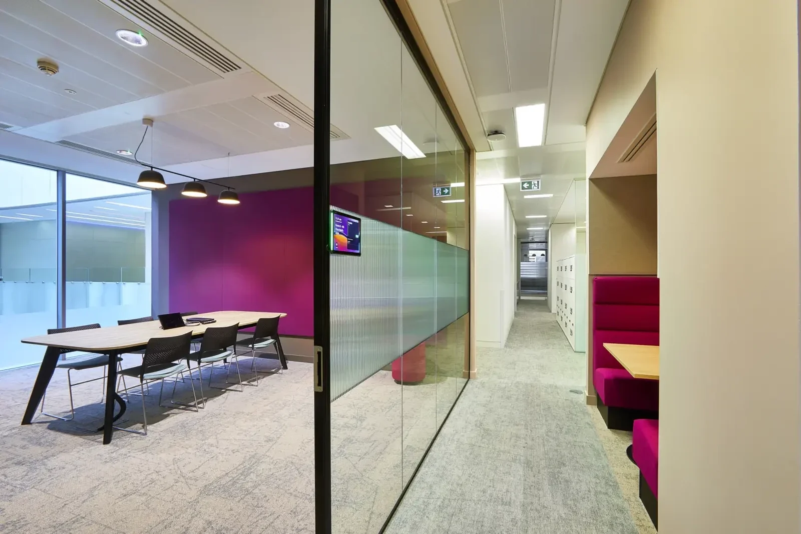Meeting room with a long timber table and magenta feature wall, seen past a glazed door, with a corridor of lockers and a magenta banquette booth beyond